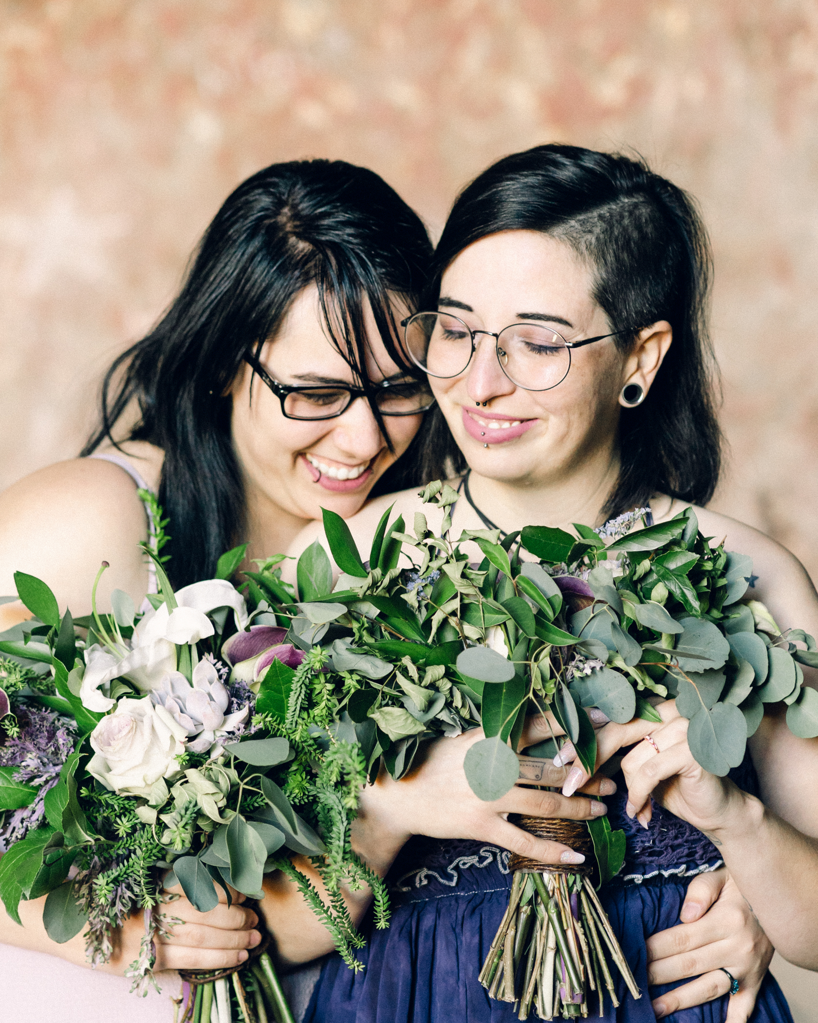 Two brides stand together with their bouquets and laugh with each other.