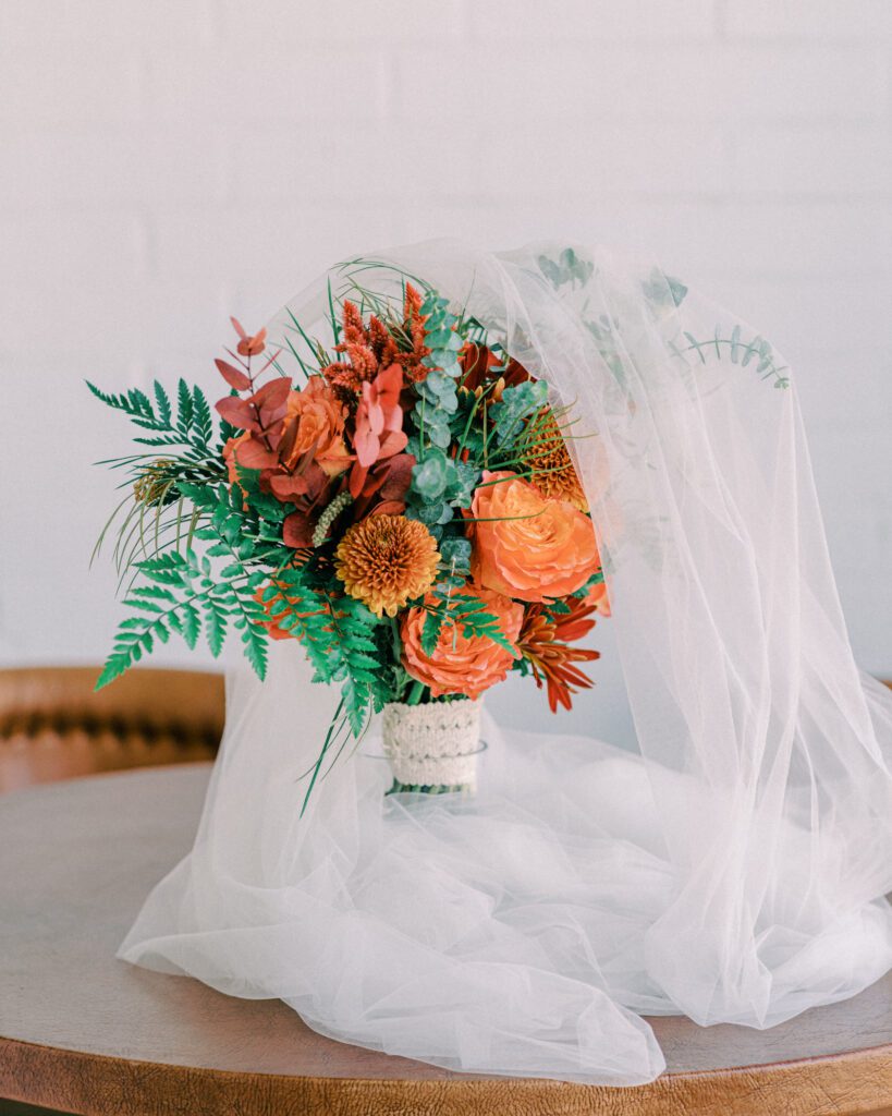 Autumn wedding bouquet with orange and red flowers wrapped in a white lace ribbon, draped with a white veil.