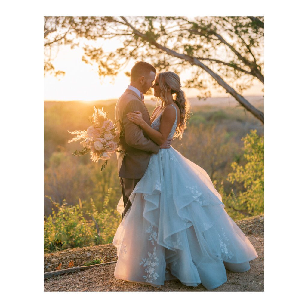 Newlywed couple embracing at sunset with a natural backdrop, bride in a lace dress and groom in a beige suit.