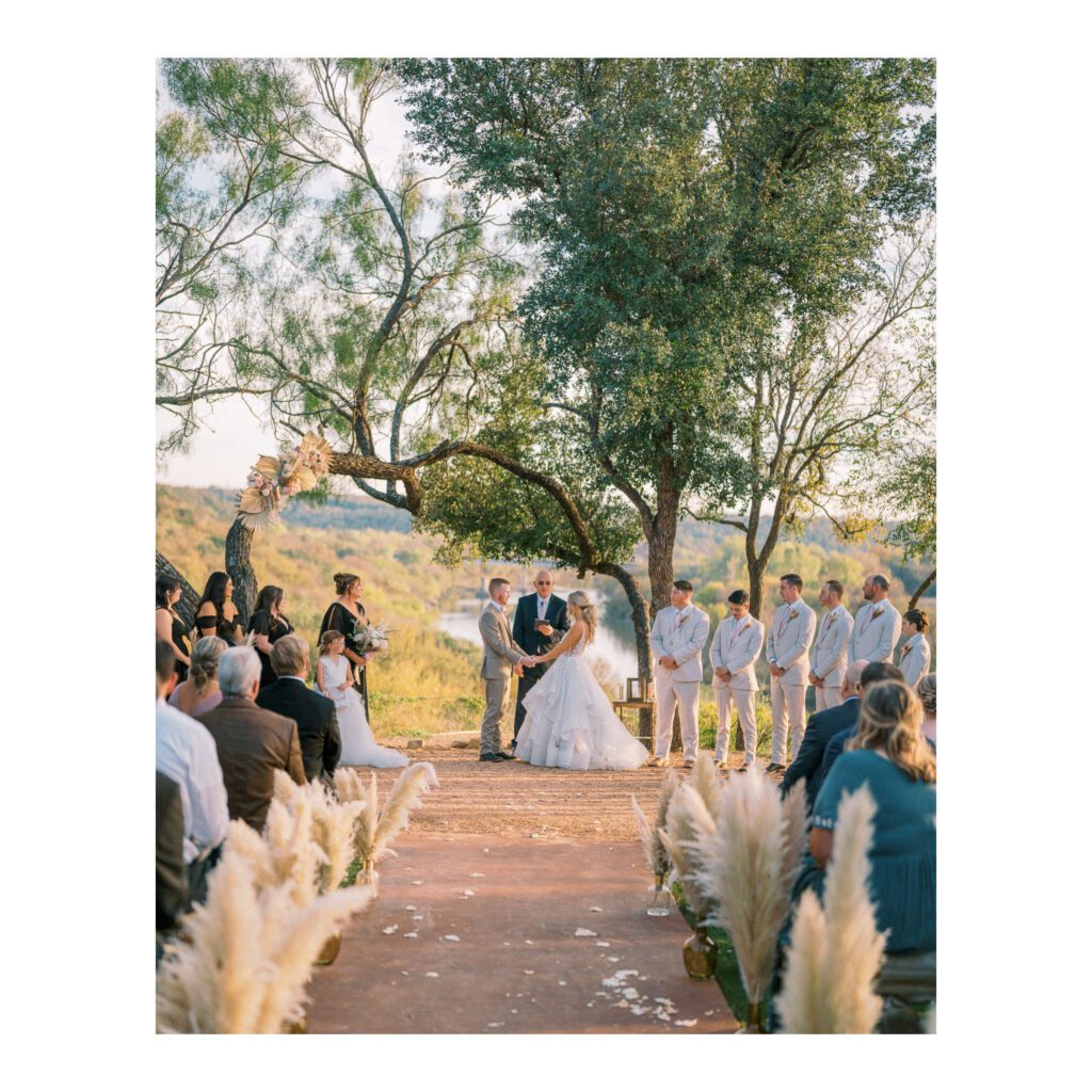 Outdoor wedding ceremony with couple exchanging vows under a tree, surrounded by guests and groomsmen in Texas.