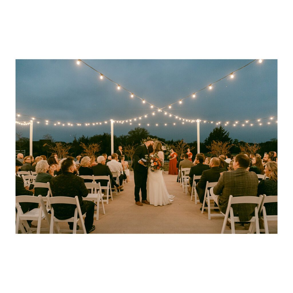 Outdoor wedding ceremony at dusk with string lights and a couple kissing at the altar surrounded by guests.